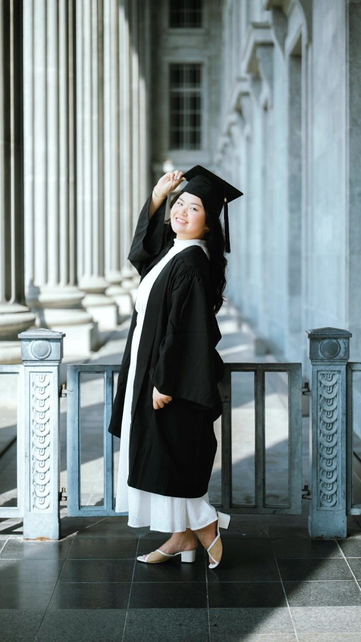 Woman in black graduation cap and gown smiling beside large stone columns.