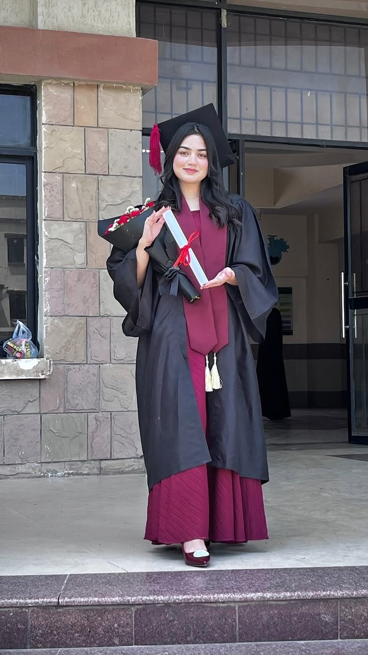 Graduating woman in black gown and cap holding a diploma and flowers outdoors.