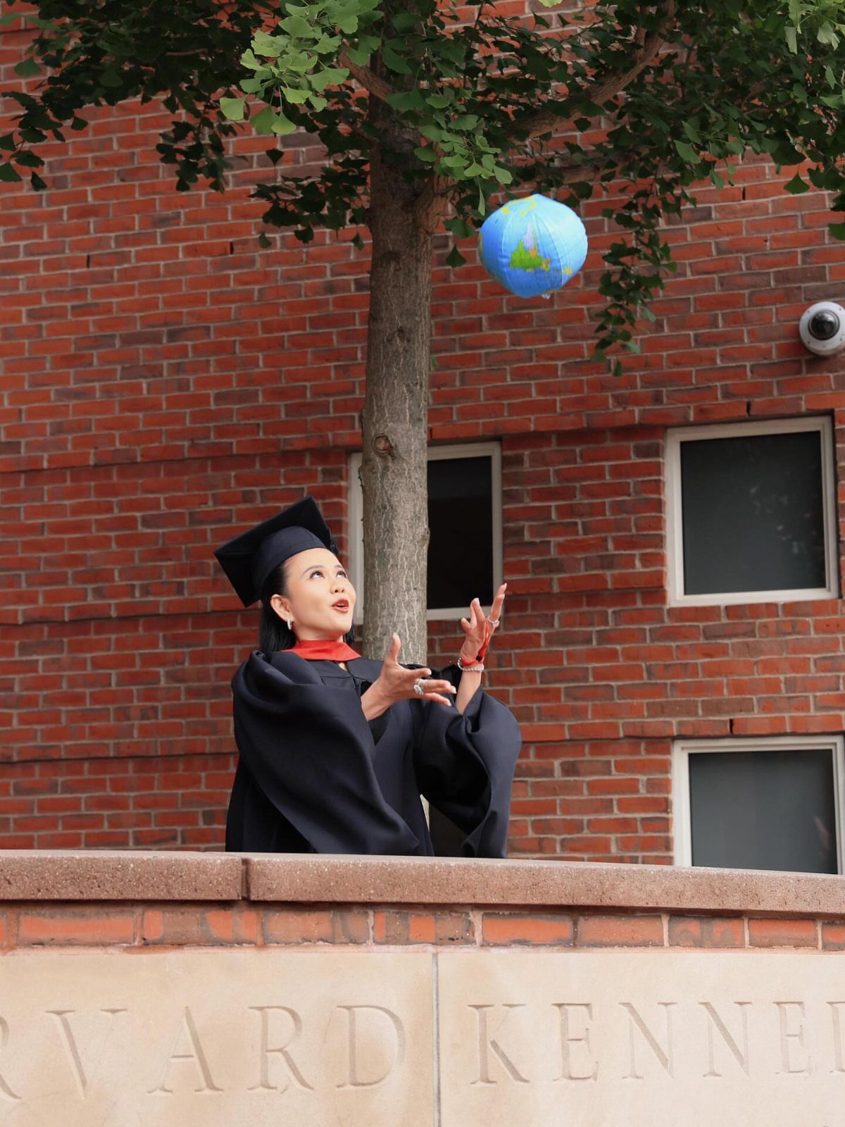 Graduate in black cap and gown tossing a globe in the air outdoors.