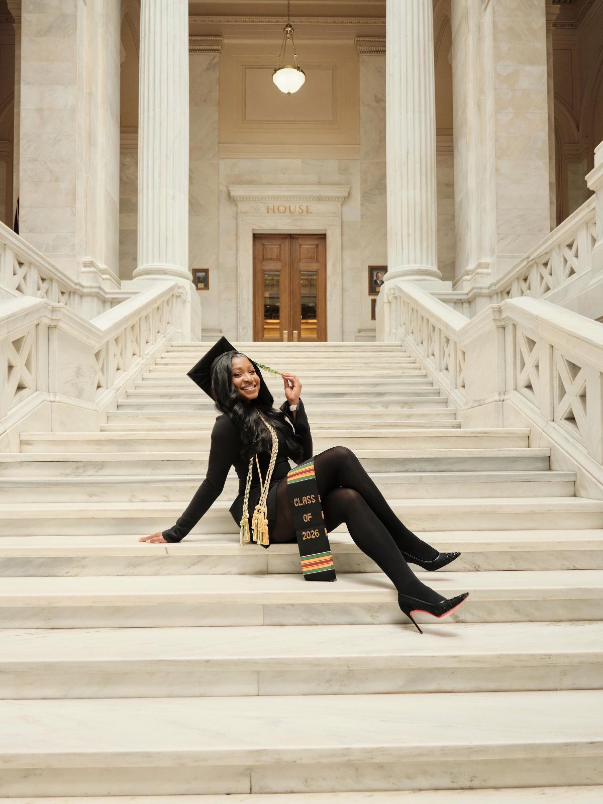 Woman in black graduation cap and gown sitting on white marble stairs.