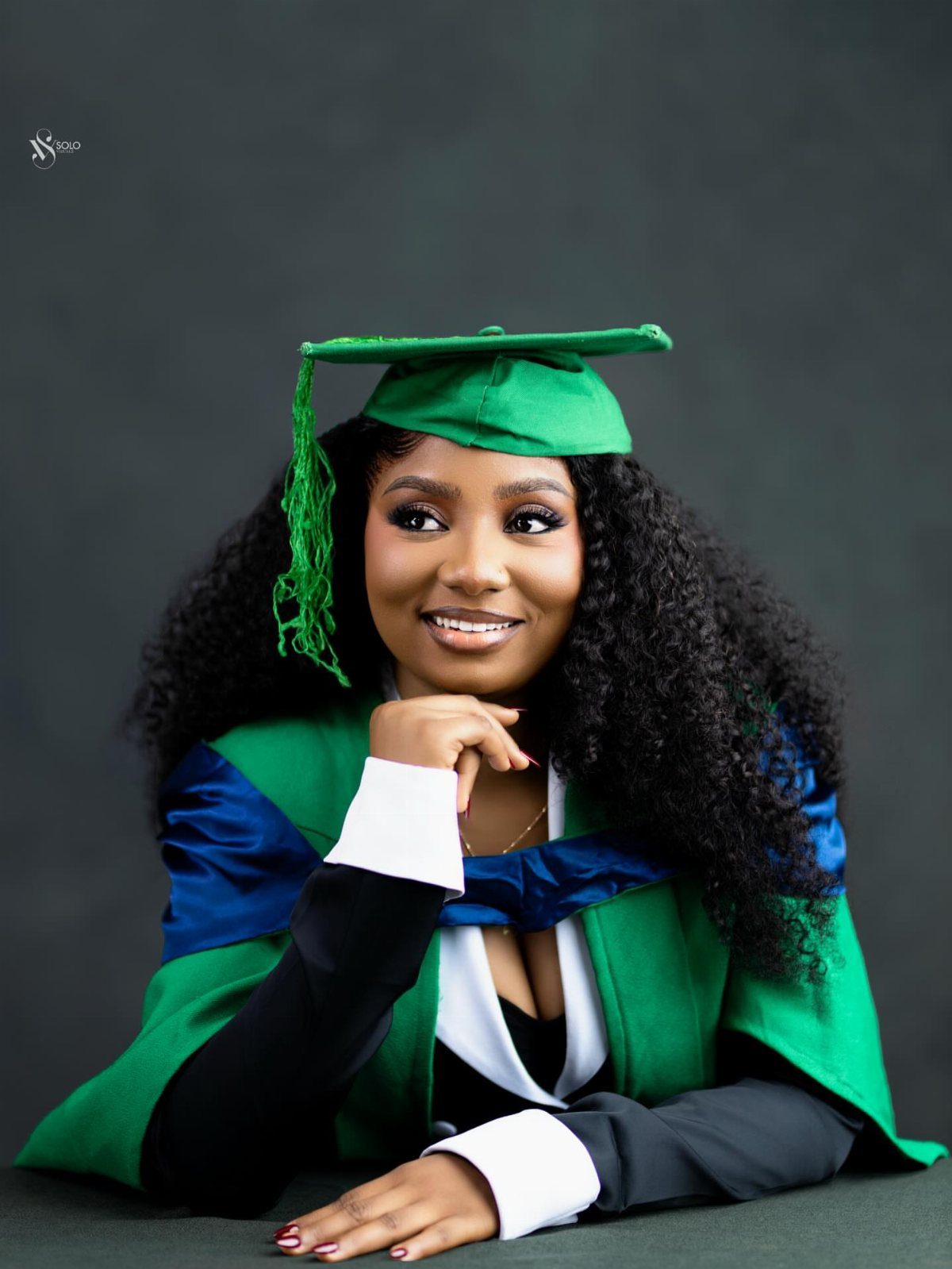 Smiling woman in green graduation cap and gown posing against a dark background