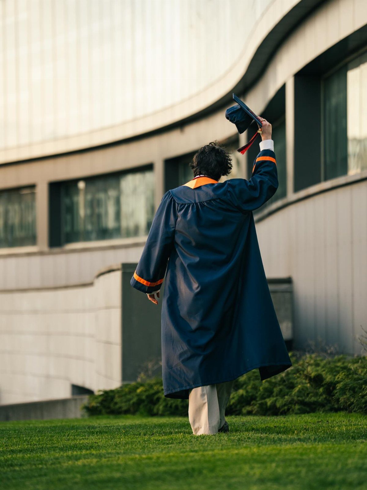 Graduate in navy gown walking away from camera toward a modern building.