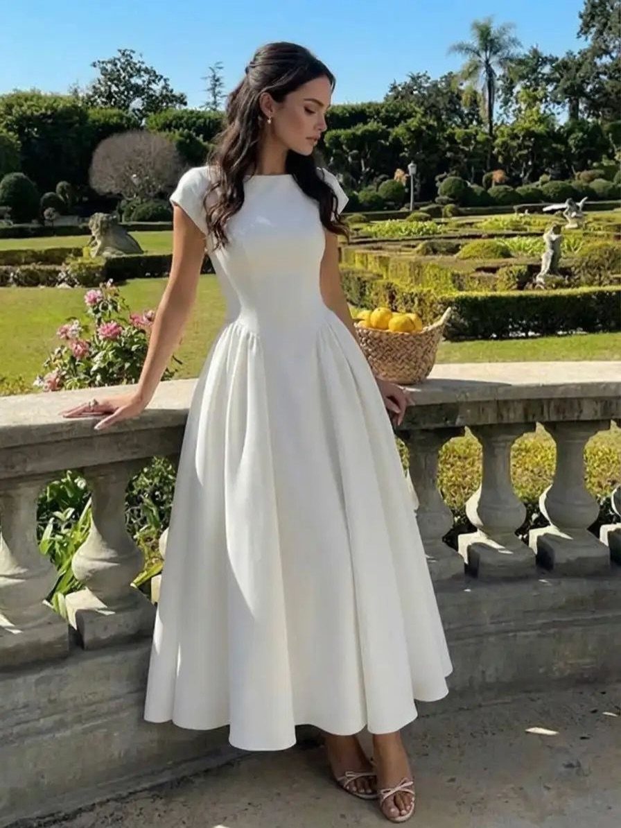 Woman in a white midi dress standing by a stone railing in a garden