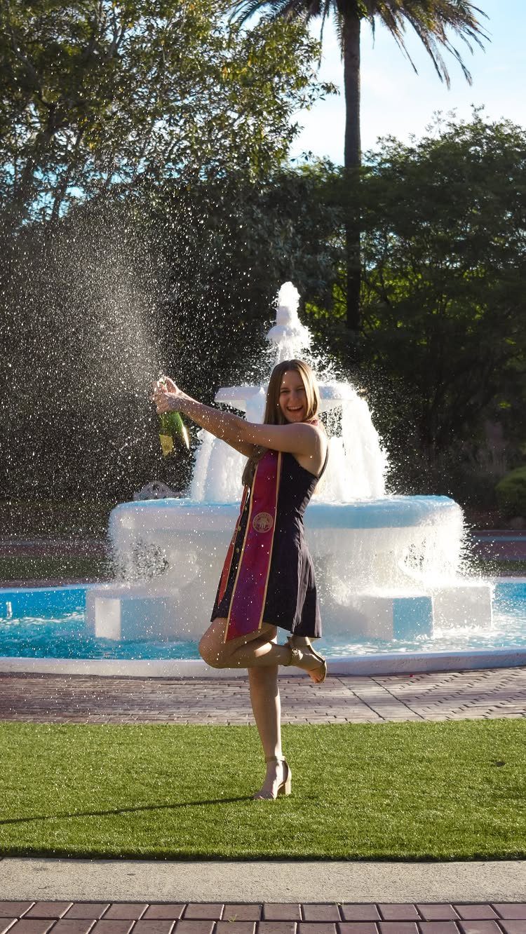 Woman in graduation stole popping a champagne bottle in front of a fountain