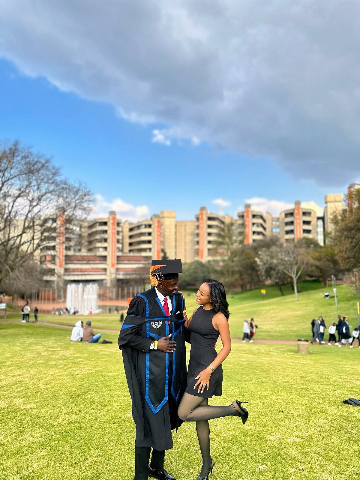Graduating man in gown and woman in black dress posing on campus lawn.