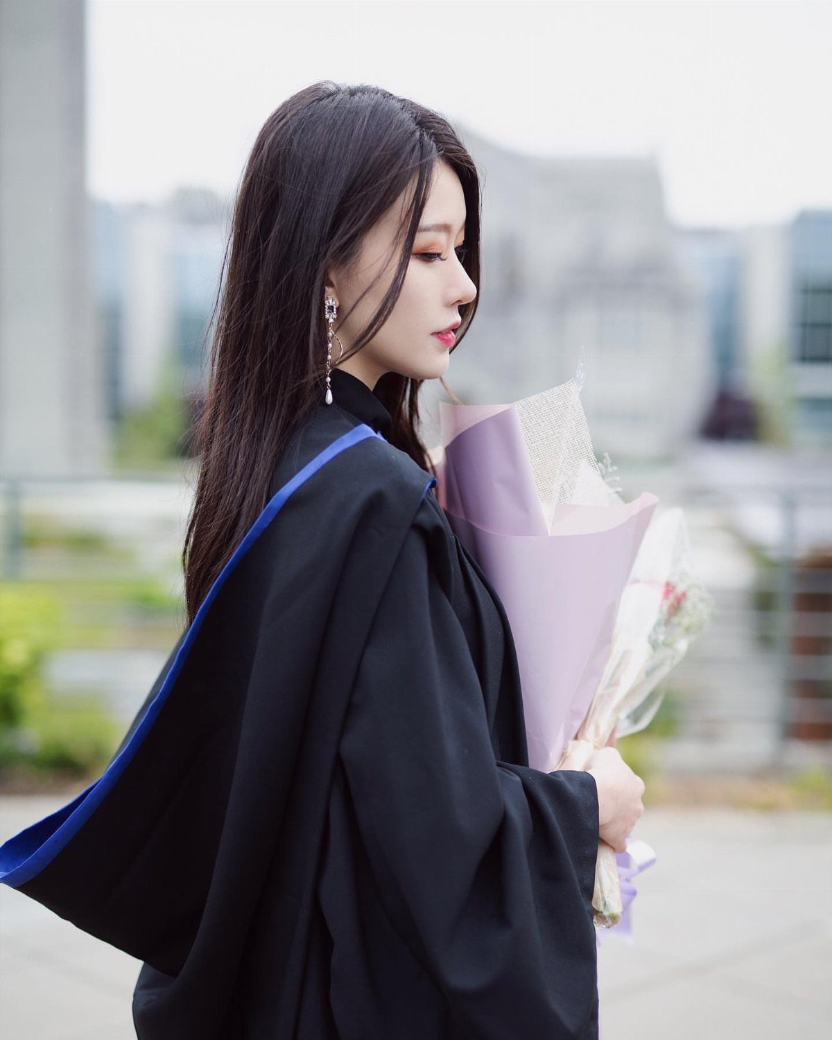 Young woman in black graduation gown holding a purple flower bouquet in profile