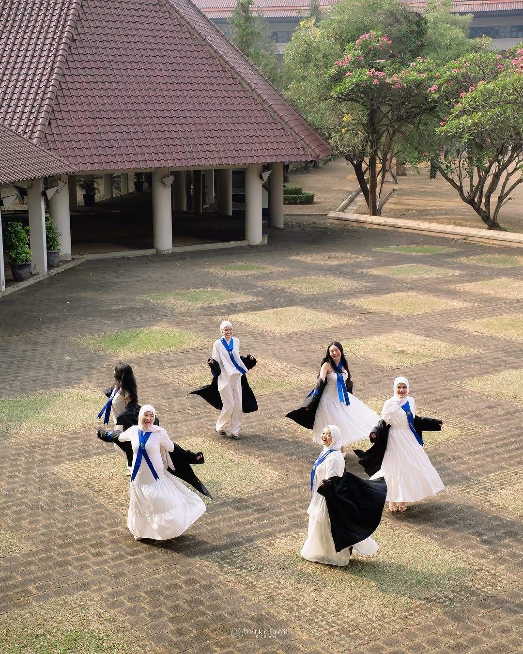 Six female graduates in white dresses and black gowns posing in a courtyard.