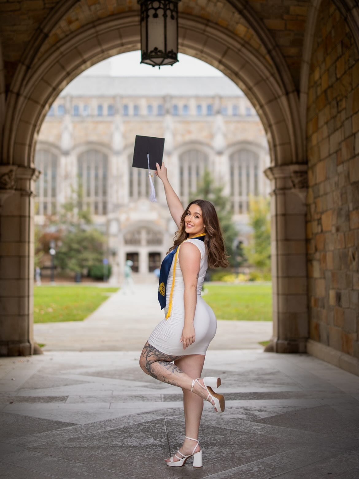 Woman in white dress holding graduation cap high under a stone archway
