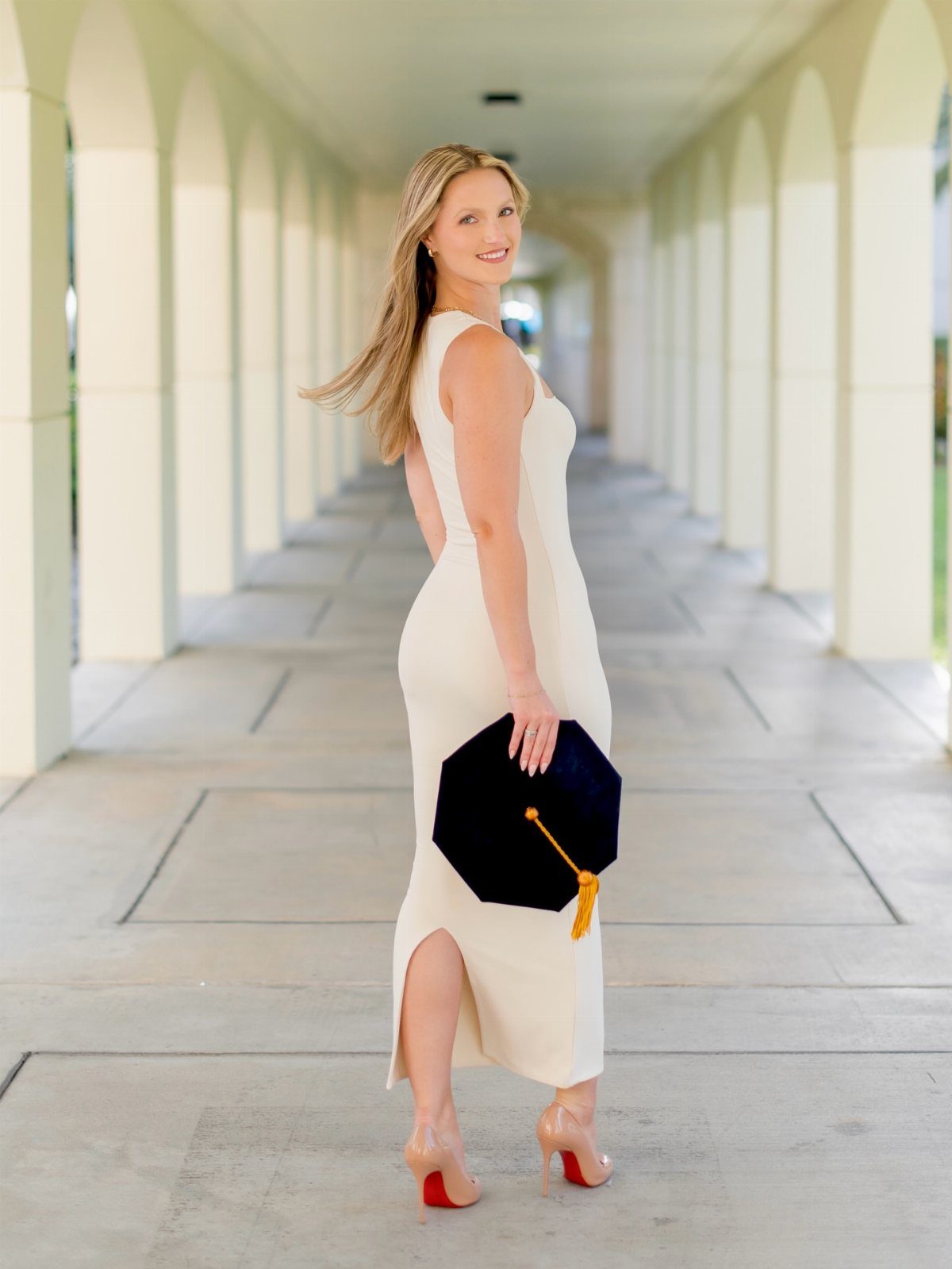 Woman in white dress holding a graduation cap in a white arched hallway