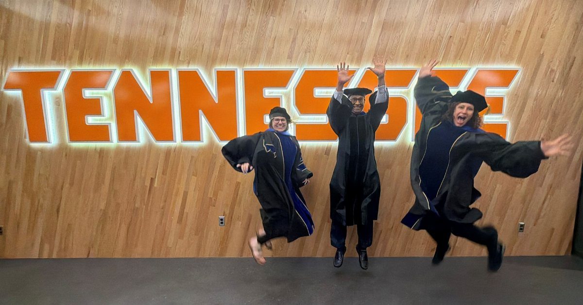 Three graduates in black gowns jumping for joy before a Tennessee sign.