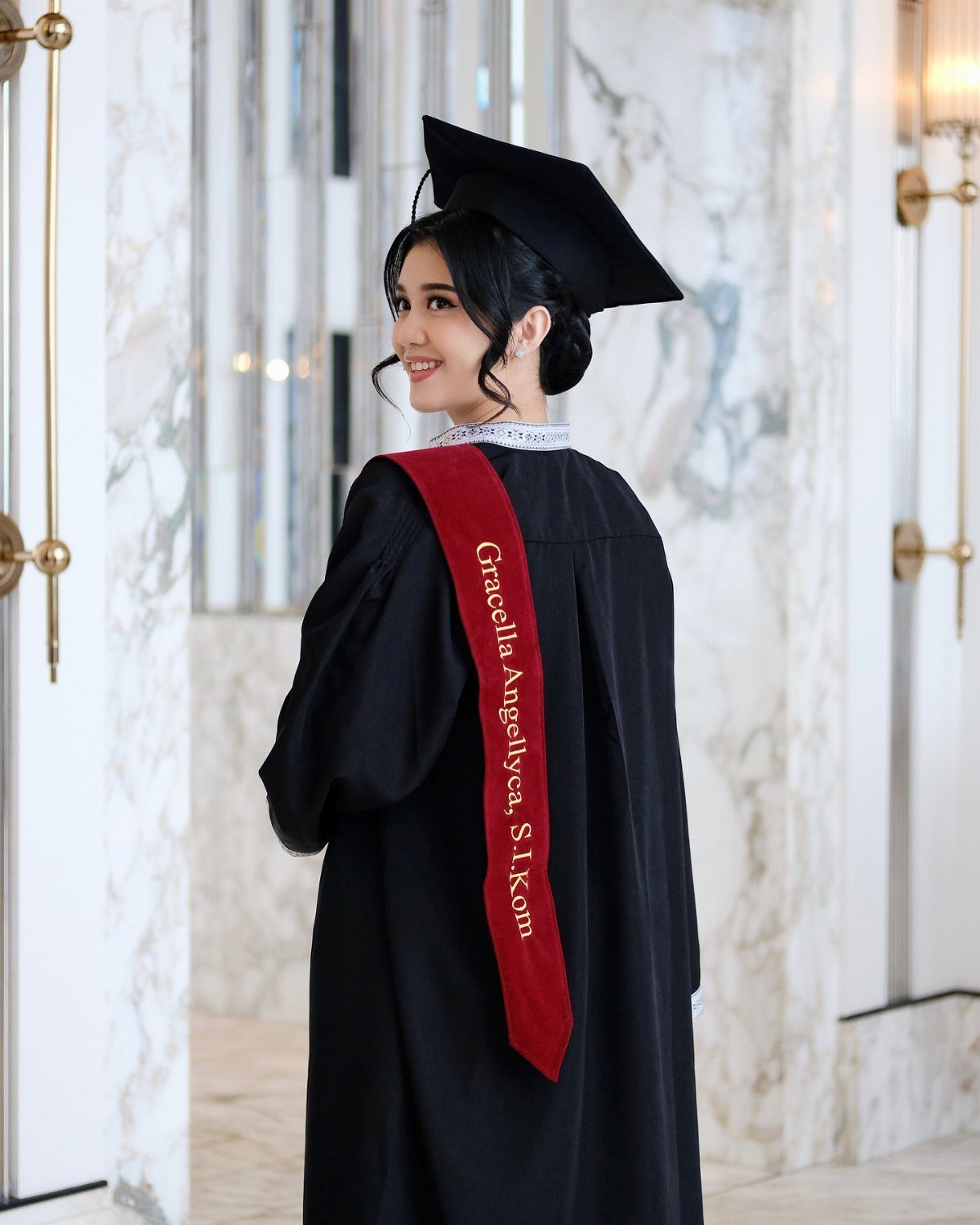 Young woman in black graduation gown and cap smiling over her shoulder indoors