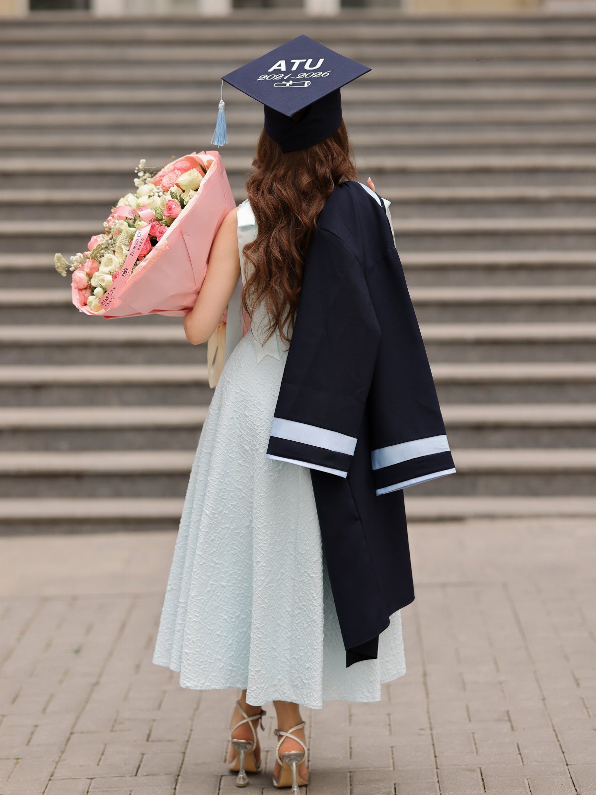 Woman in blue dress and graduation cap walking away toward stone steps