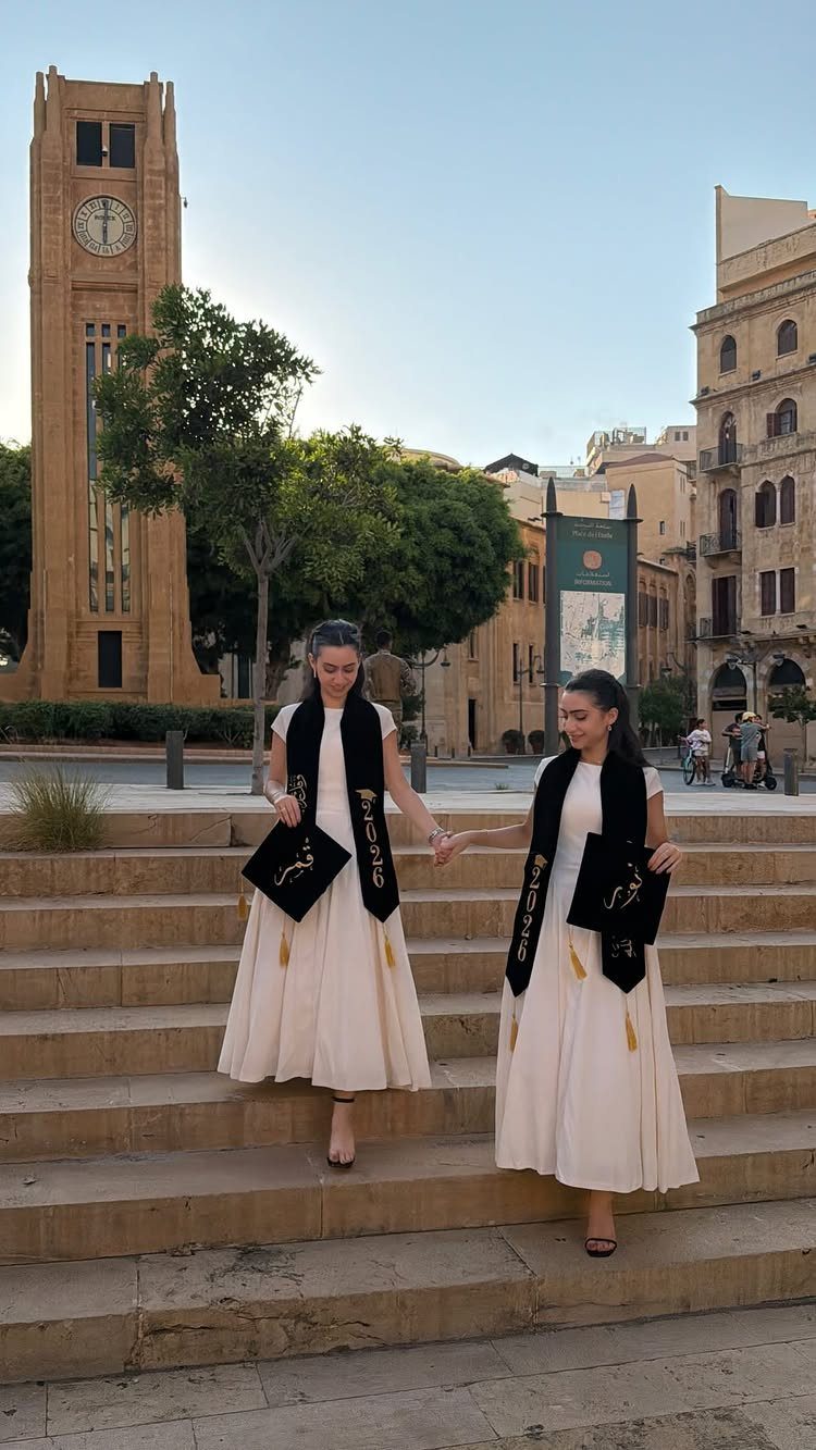 Two female graduates in cream dresses and black stoles walking down stone steps.