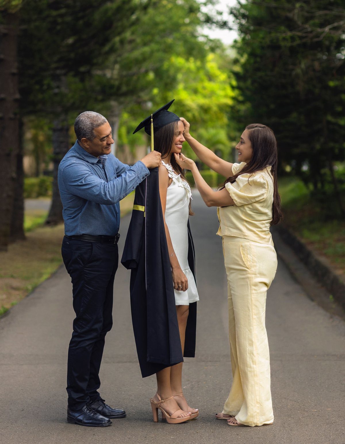 Graduating woman in white dress being helped with her cap and gown by parents.