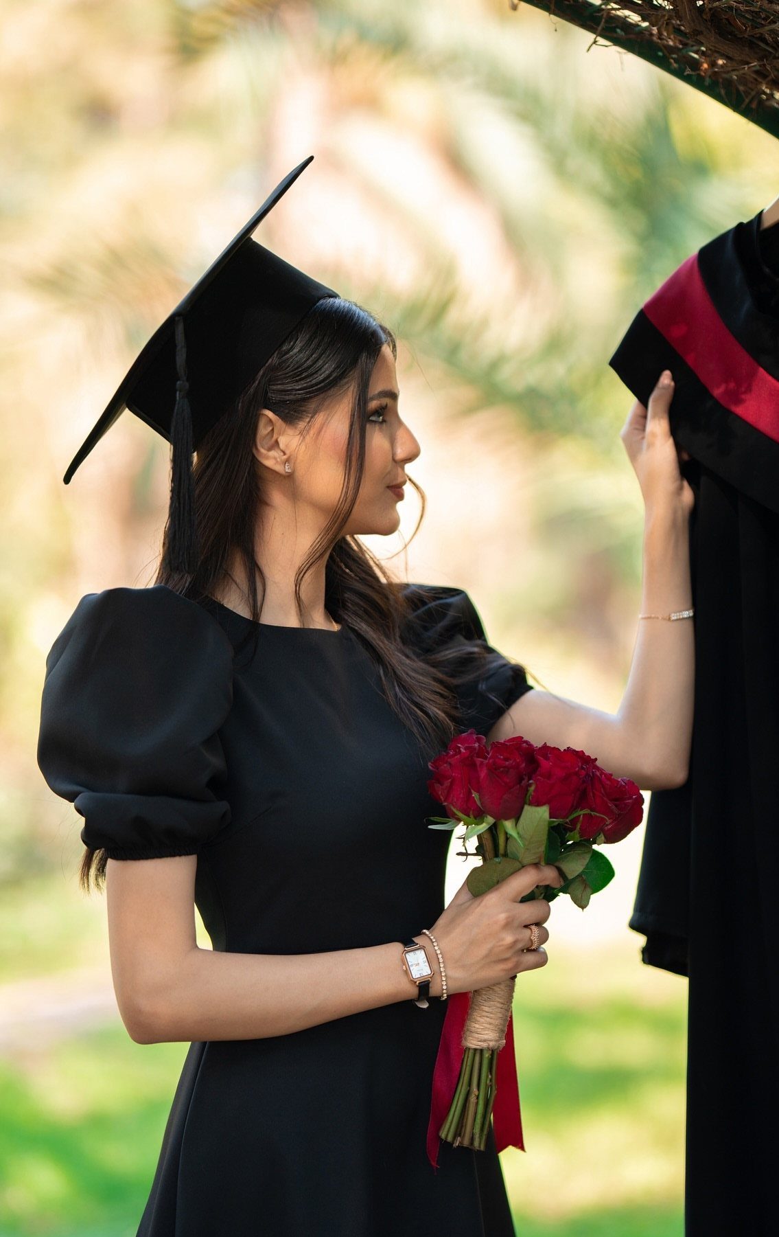 Woman in graduation cap holding red roses and touching a graduation gown outdoors.