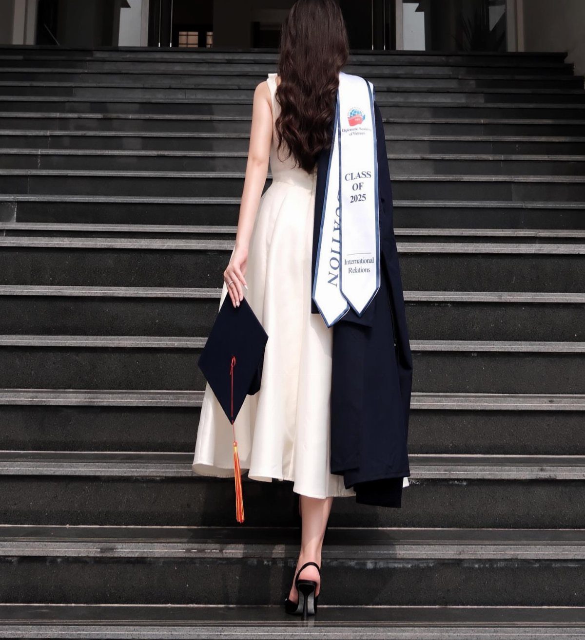 Woman in white dress walking up stairs carrying graduation cap and gown
