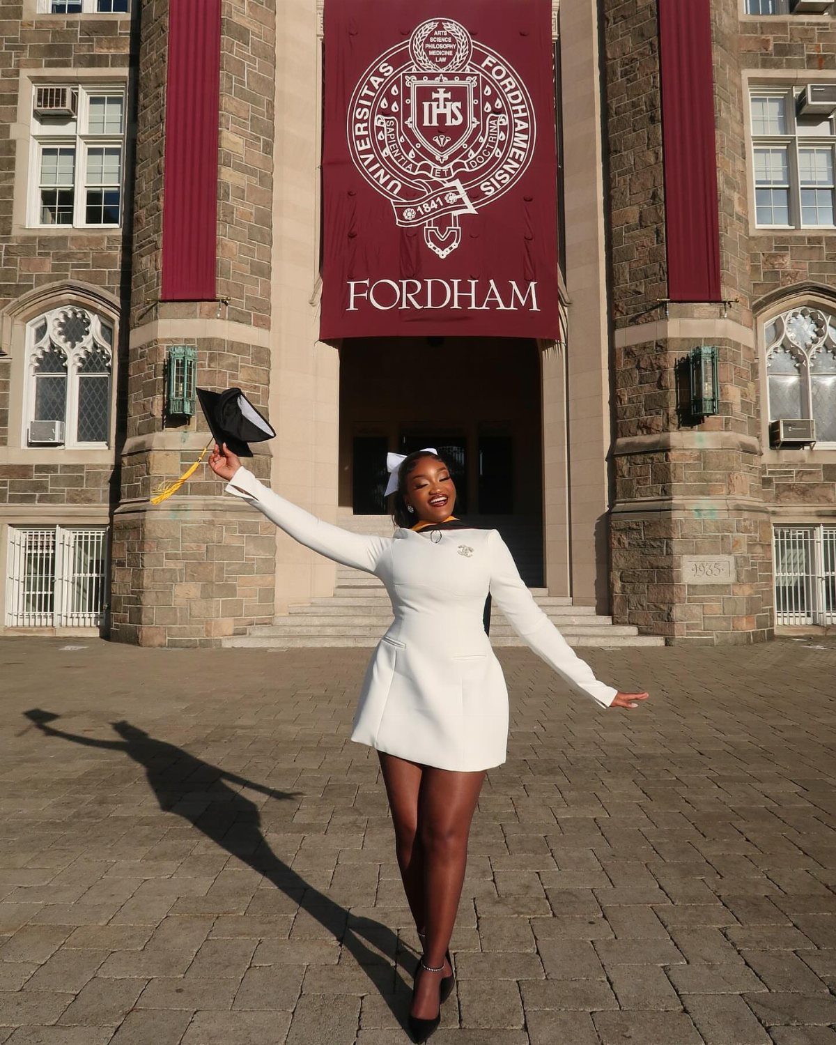 Woman in a white dress holding a graduation cap before a university building