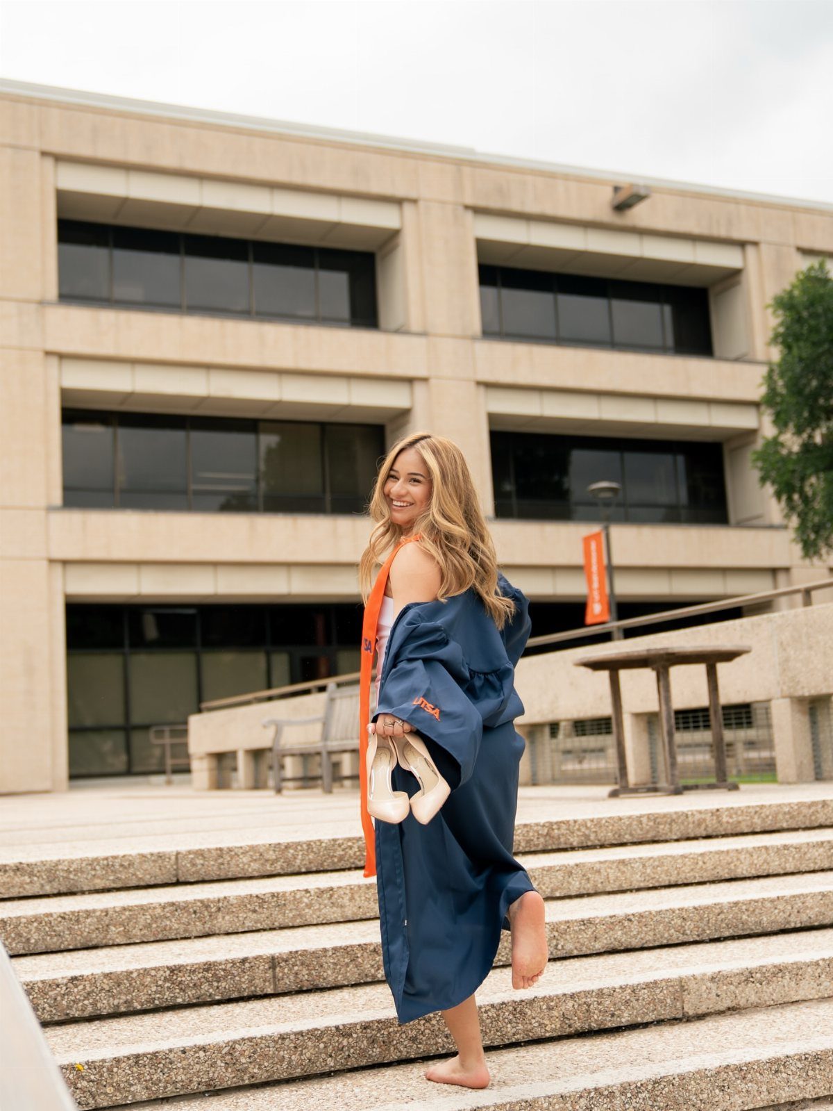 Smiling graduate in navy gown and orange stole standing barefoot on concrete stairs