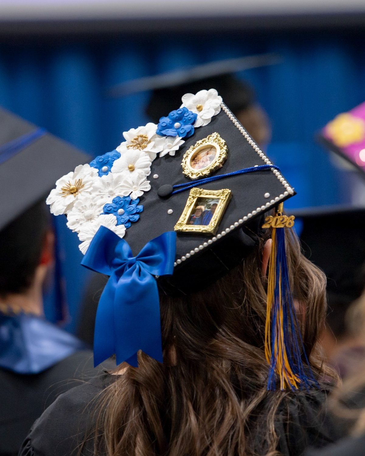 Decorated graduation cap with blue and white flowers, small photos, and a bow.