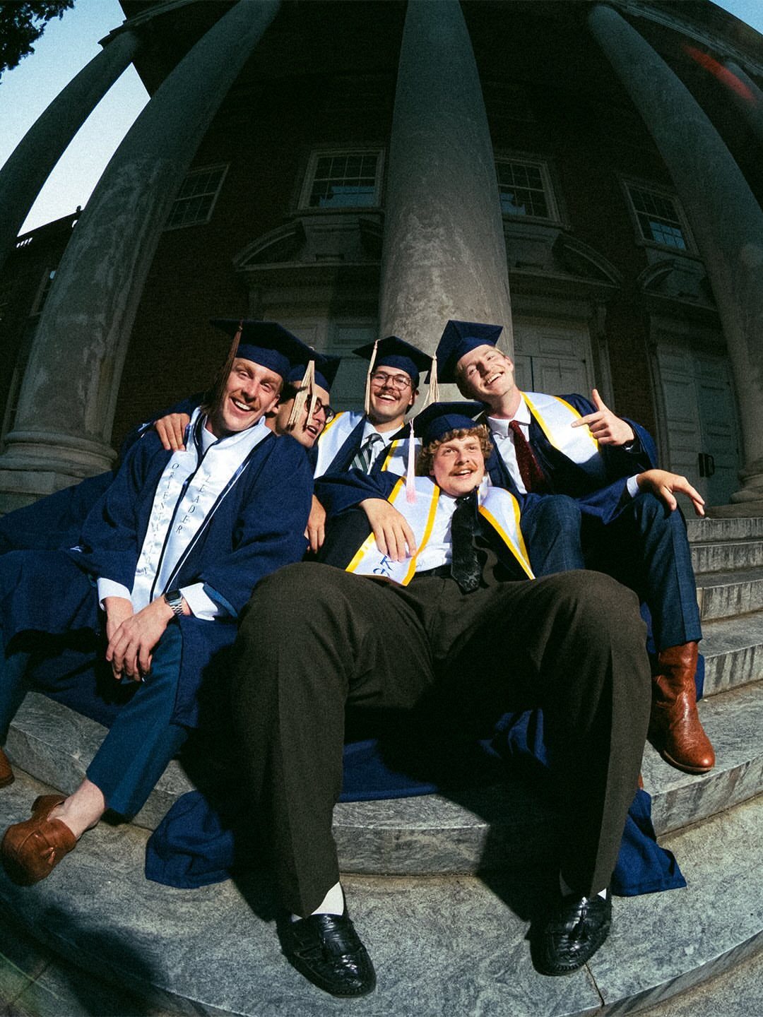 Five graduates in blue gowns and caps posing on stone steps by columns.