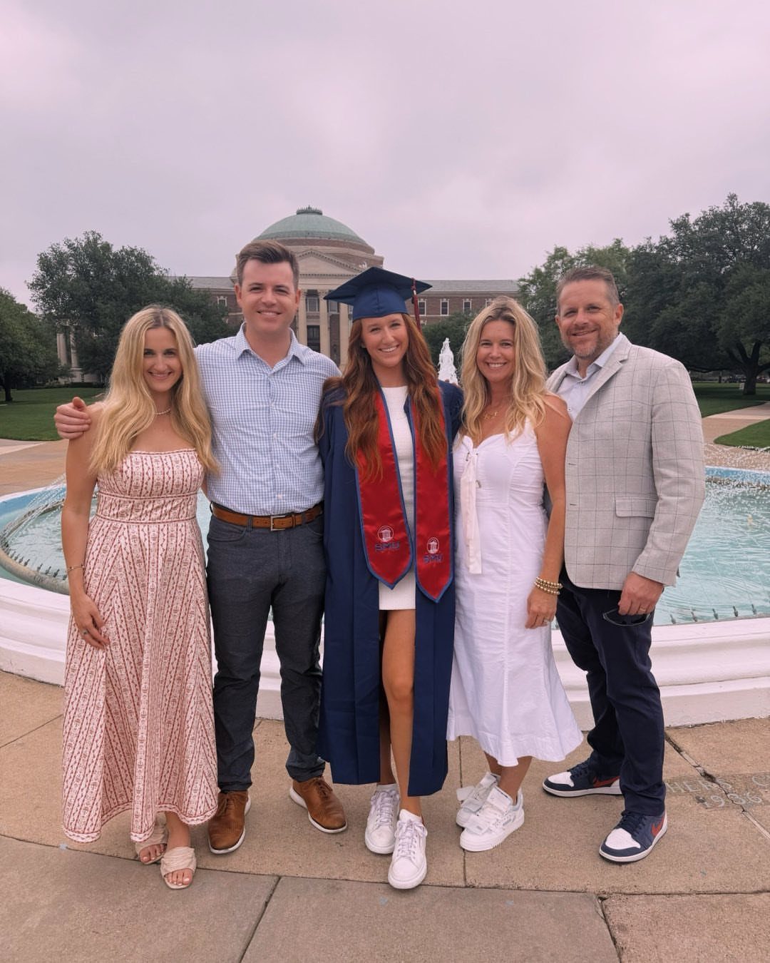 Graduating woman in blue gown posing with four family members by a campus fountain.