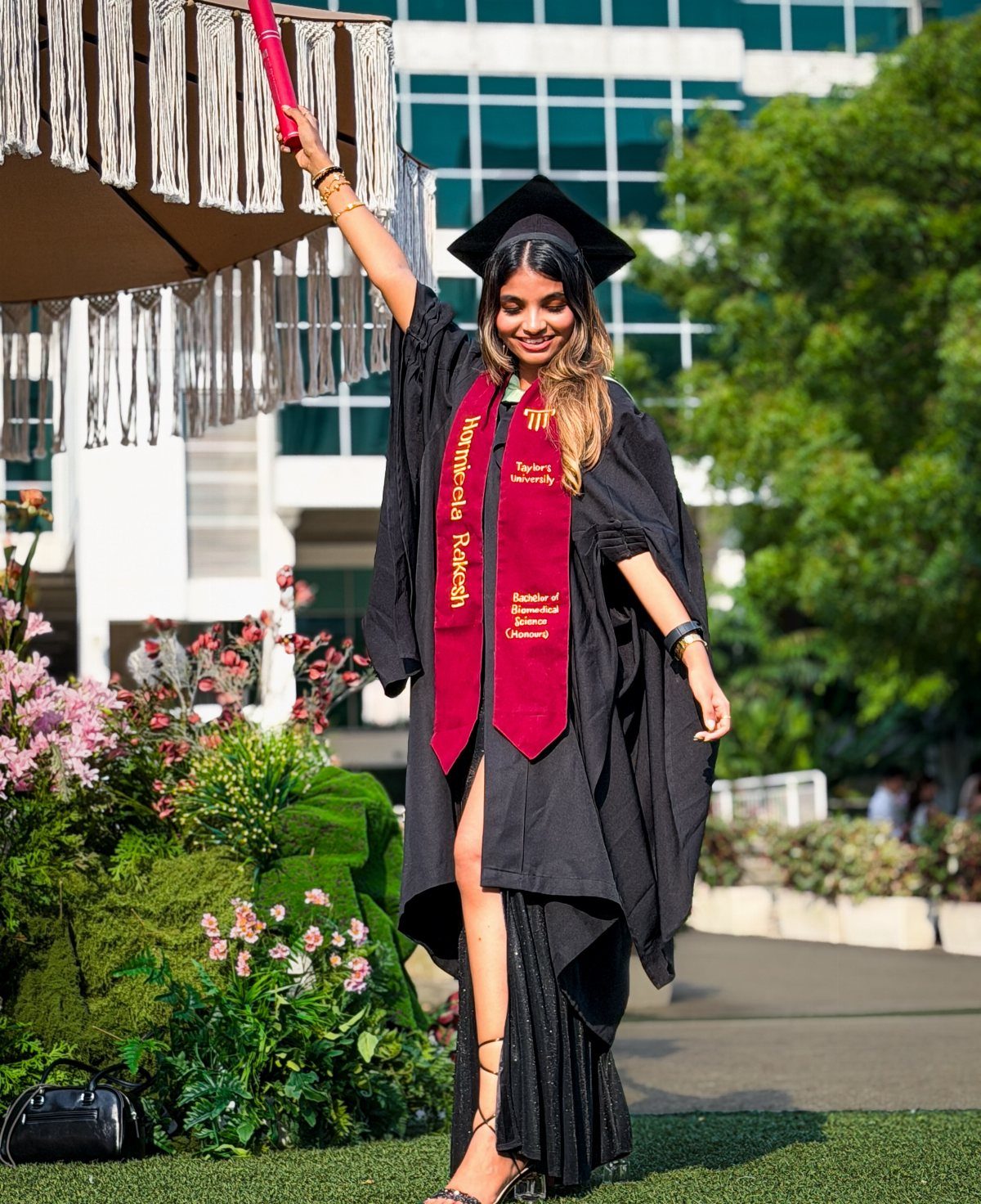 Graduate in black gown and red stole holding a diploma tube high outdoors.