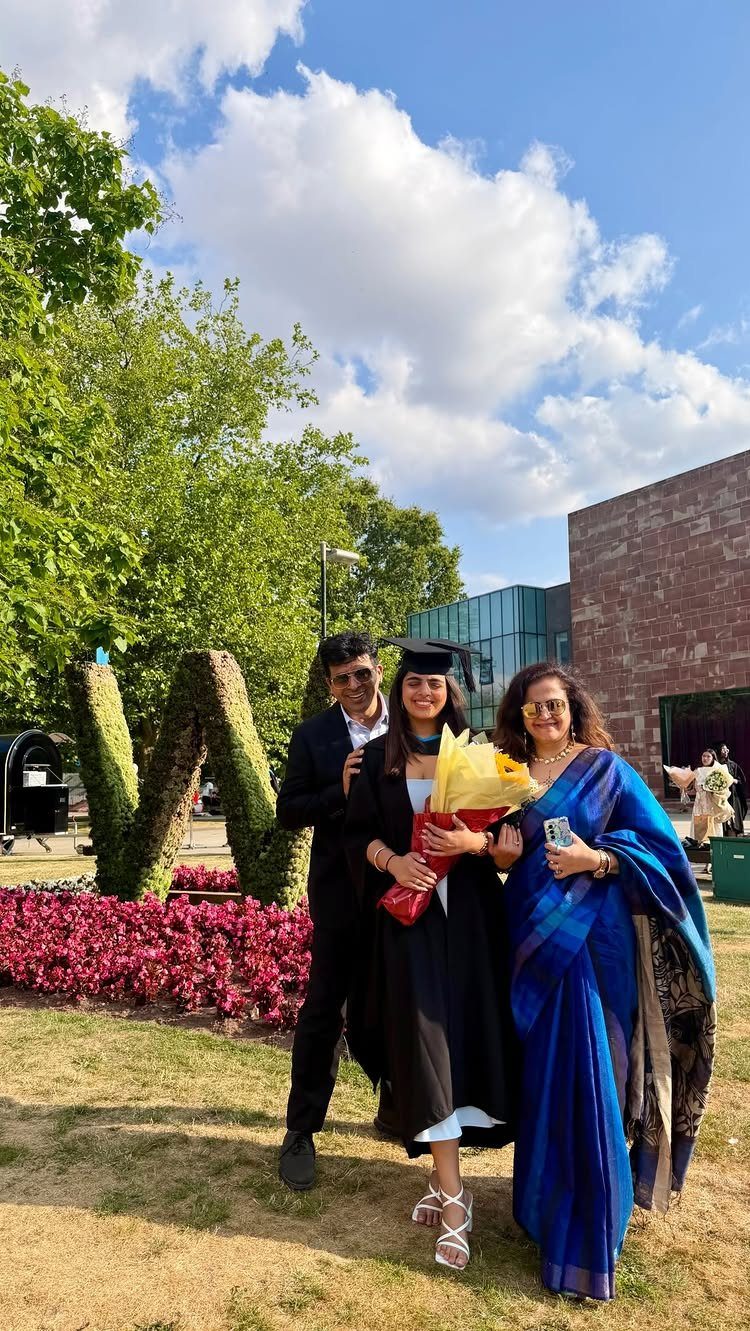 Graduating woman in cap and gown posing with parents on a sunny campus lawn