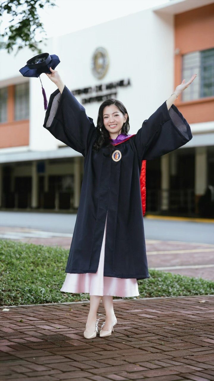 Graduating woman in navy gown and pink skirt holding her cap aloft outdoors