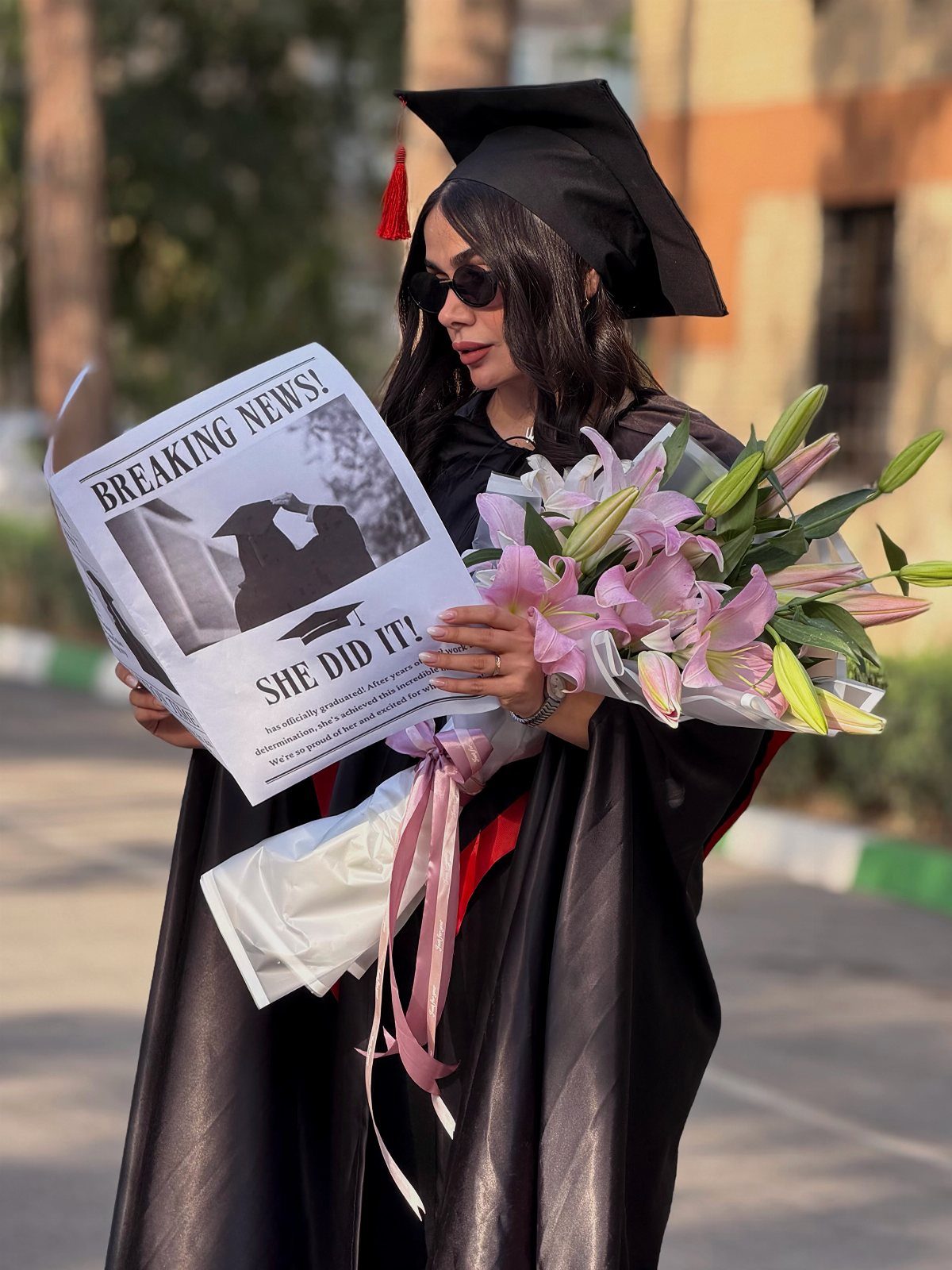 Graduating woman in black gown holding pink lilies and a custom newspaper announcement.