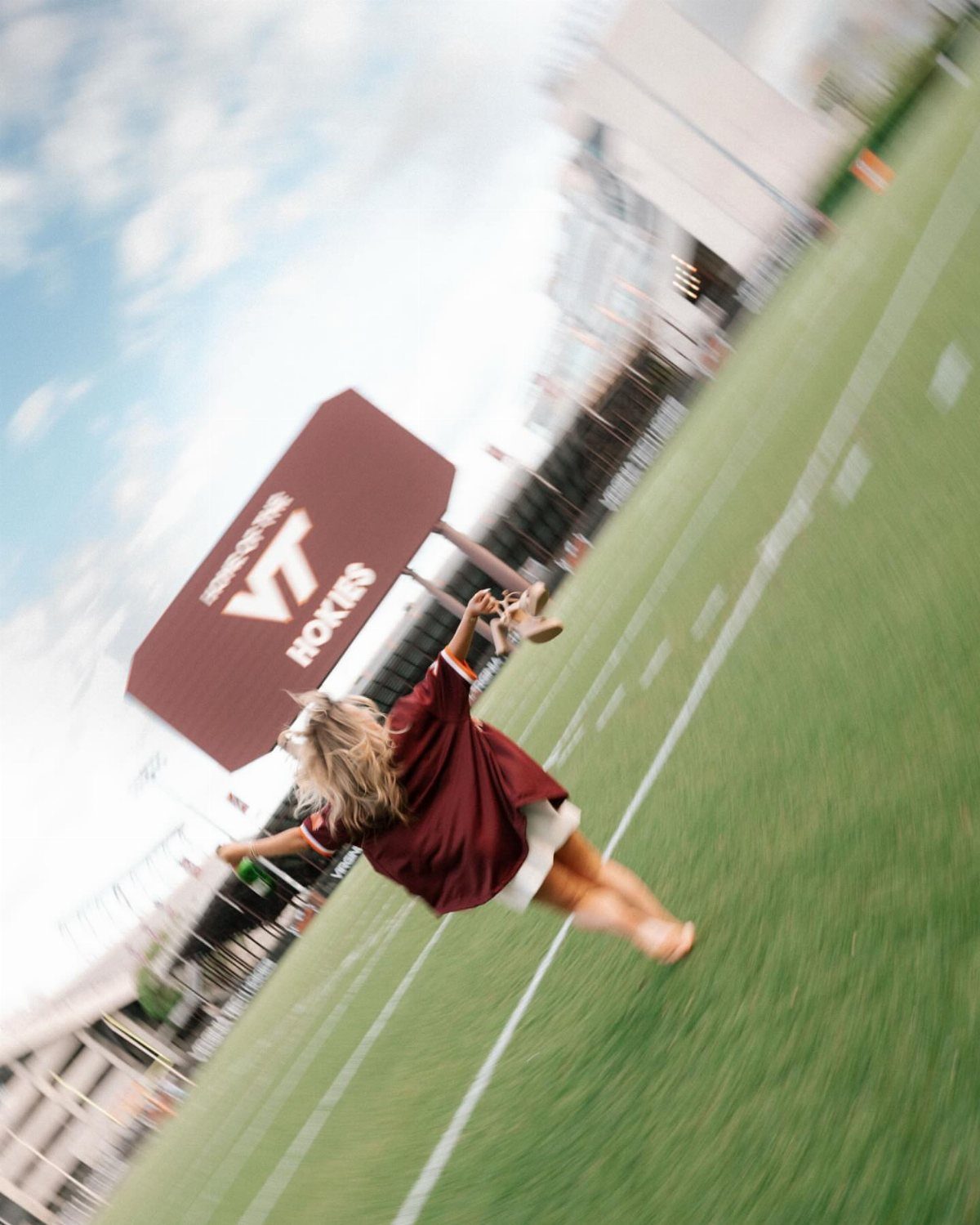 Woman in maroon graduation gown jumping on a green athletic field outdoors