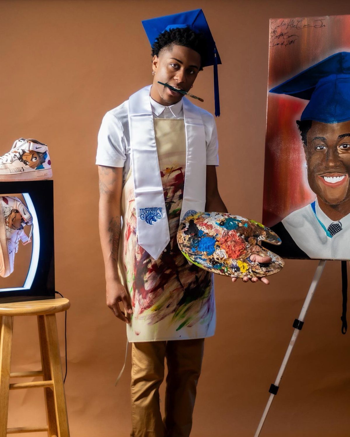 Graduating student in an apron holding a paint palette next to his portrait.