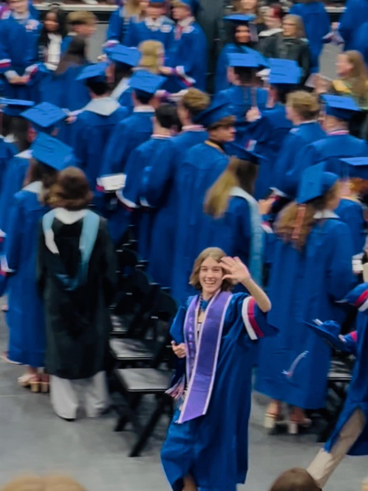 Smiling graduate in blue gown waving during a graduation ceremony with other students.
