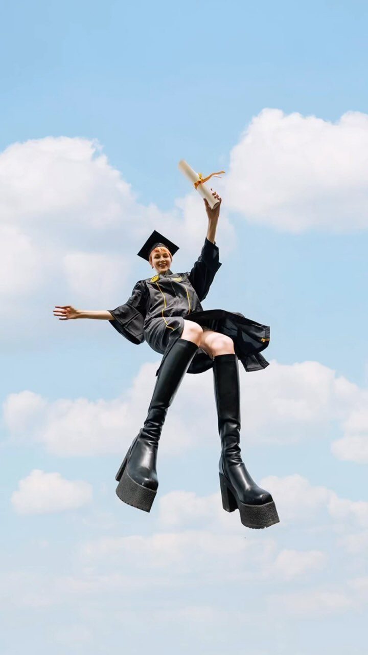 Graduate in cap, gown, and platform boots jumping against a cloudy blue sky