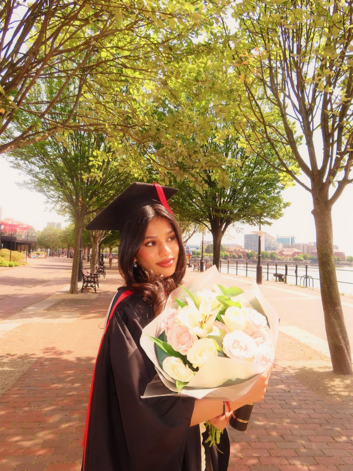 Graduate in cap and gown holding white roses during warm golden hour lighting.