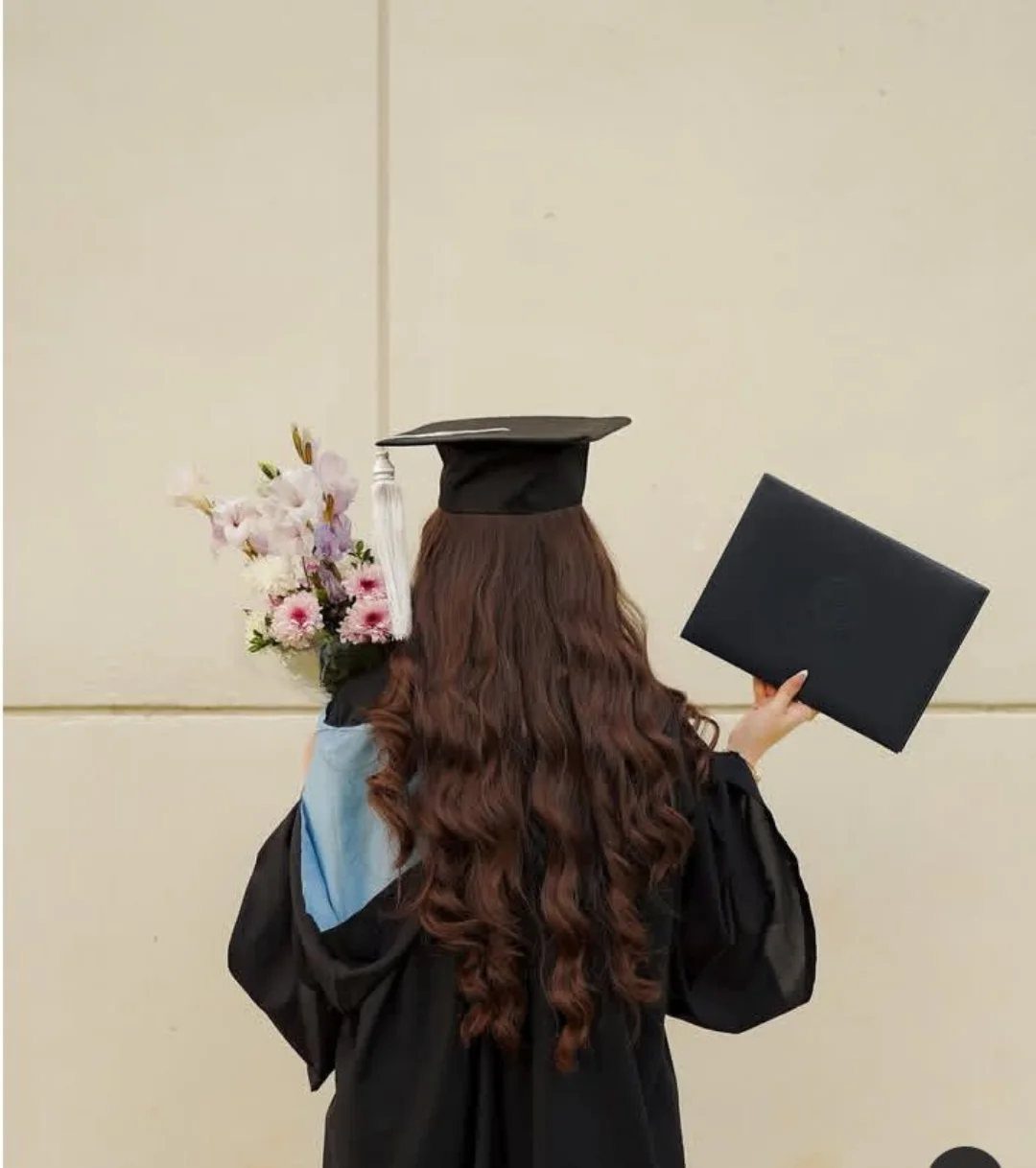 Woman in graduation gown and cap holding a diploma and flowers from behind