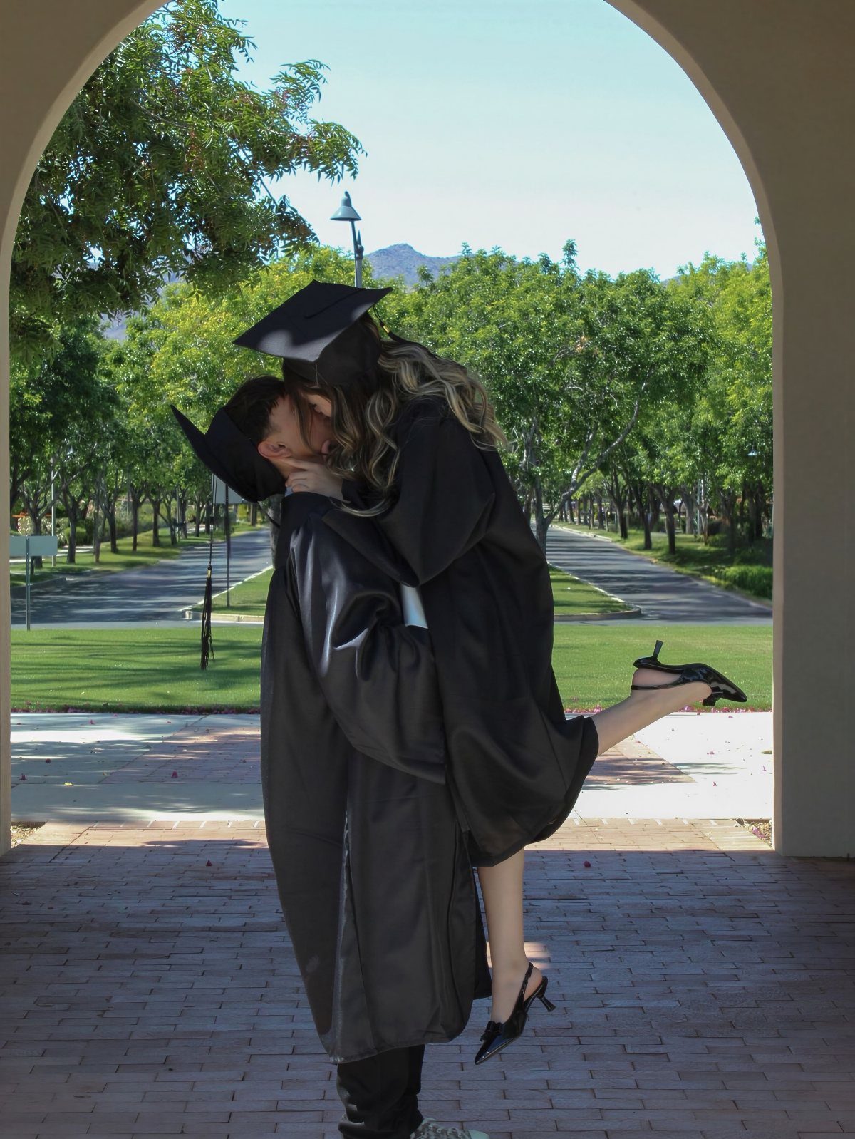 Two graduates in black caps and gowns kissing under a stone archway outdoors.
