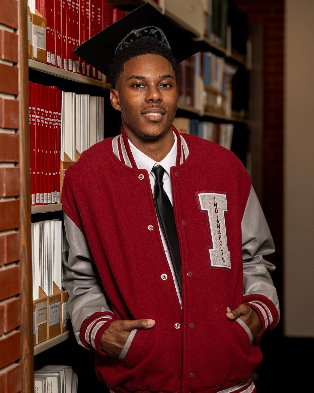 Young man in graduation cap and red letterman jacket posing in a library