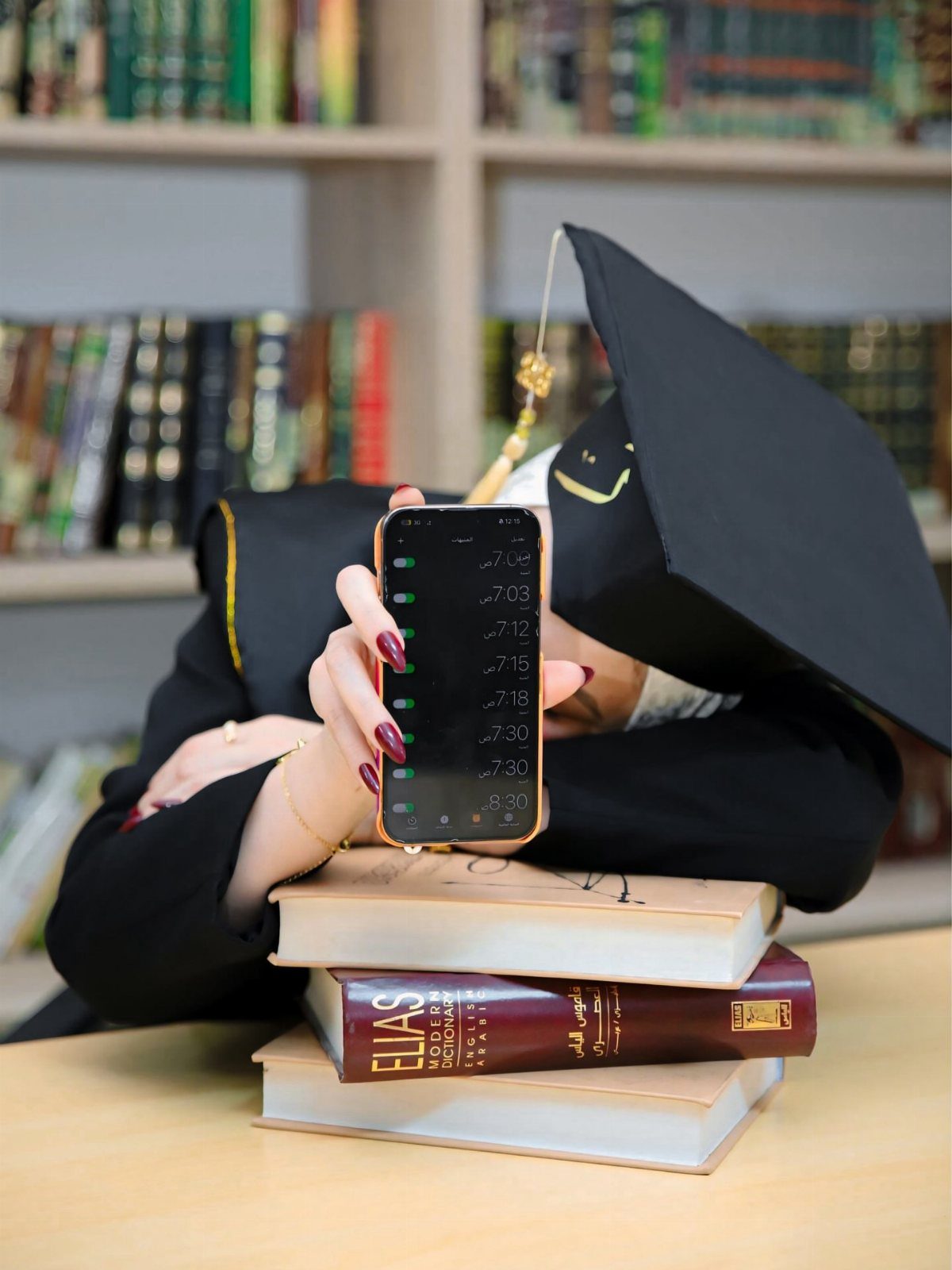 Graduate in cap and gown resting head on books in a library setting