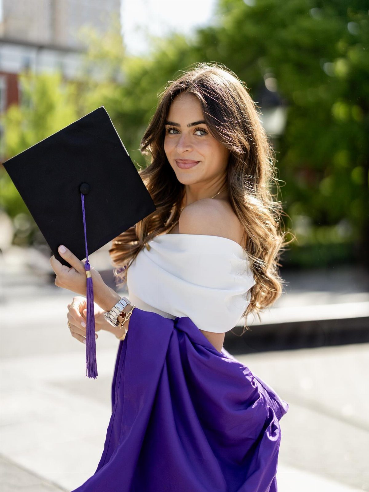 Young woman holding a graduation cap and purple gown while wearing a white top.