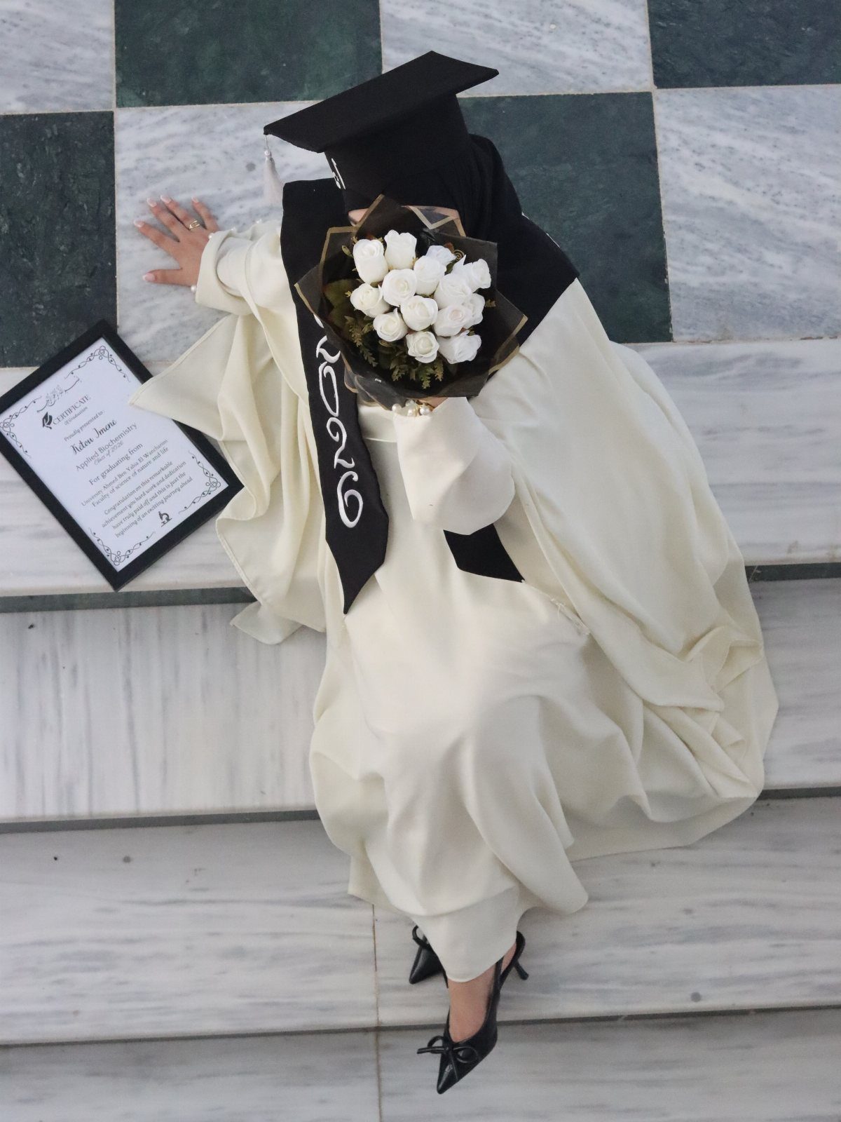 Graduate in white gown sitting on marble stairs with roses and framed diploma