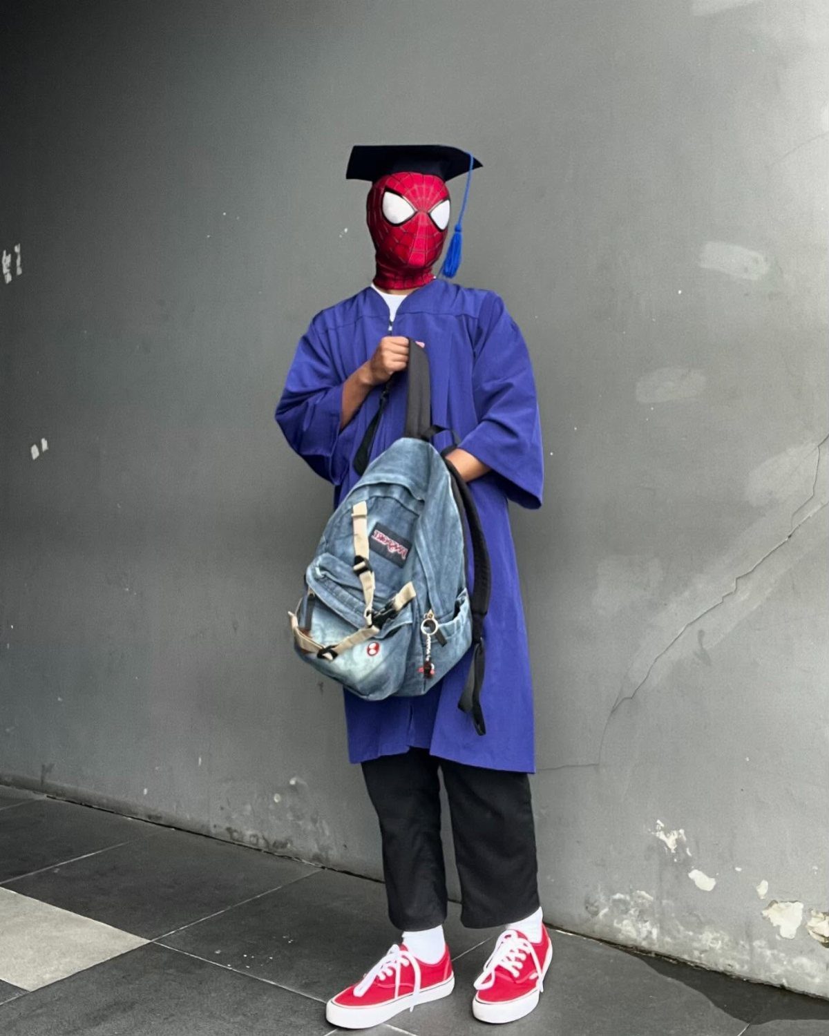 Person in Spider-Man mask and blue graduation gown holding a denim backpack.