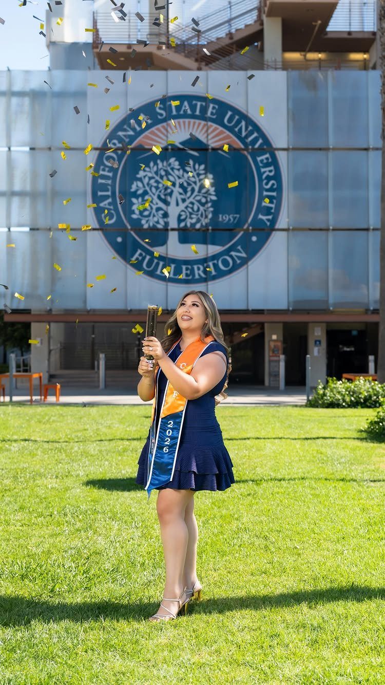 Woman in graduation attire holding a diploma with confetti falling on campus.
