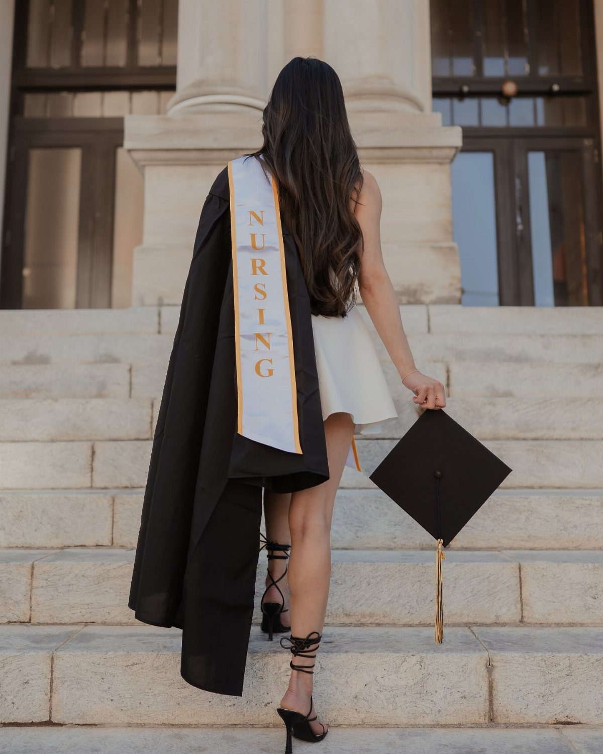 Woman in graduation attire walking up stone stairs away from the camera