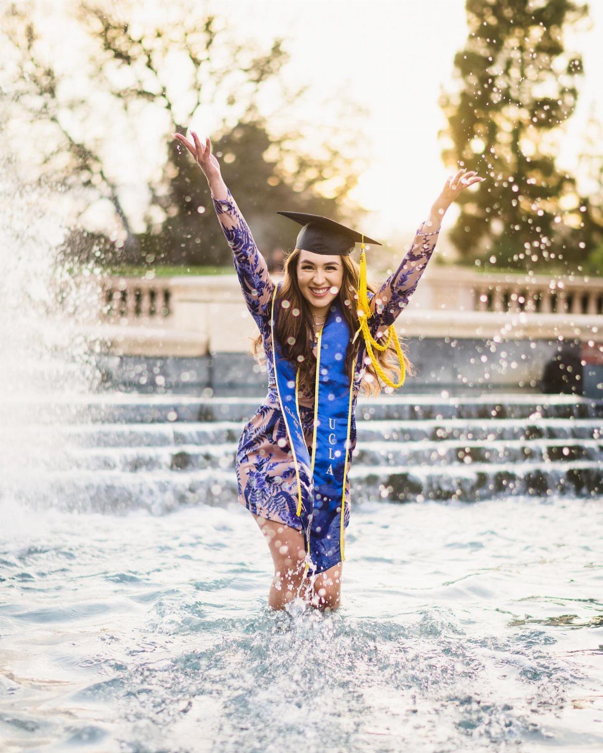 Graduating woman in a blue dress and cap smiling in a splashing fountain