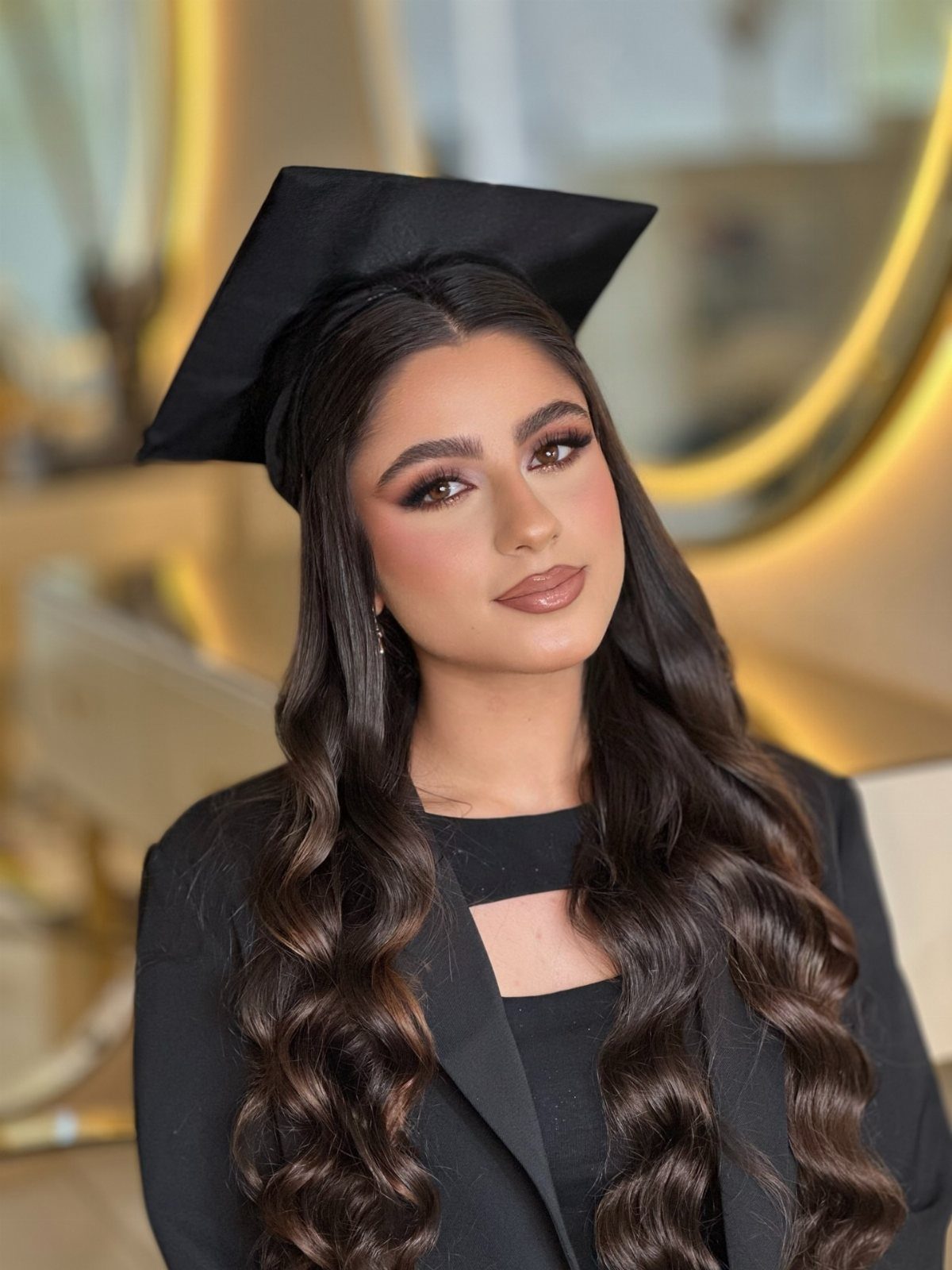 Woman in graduation cap with styled curly hair and professional makeup posing indoors.