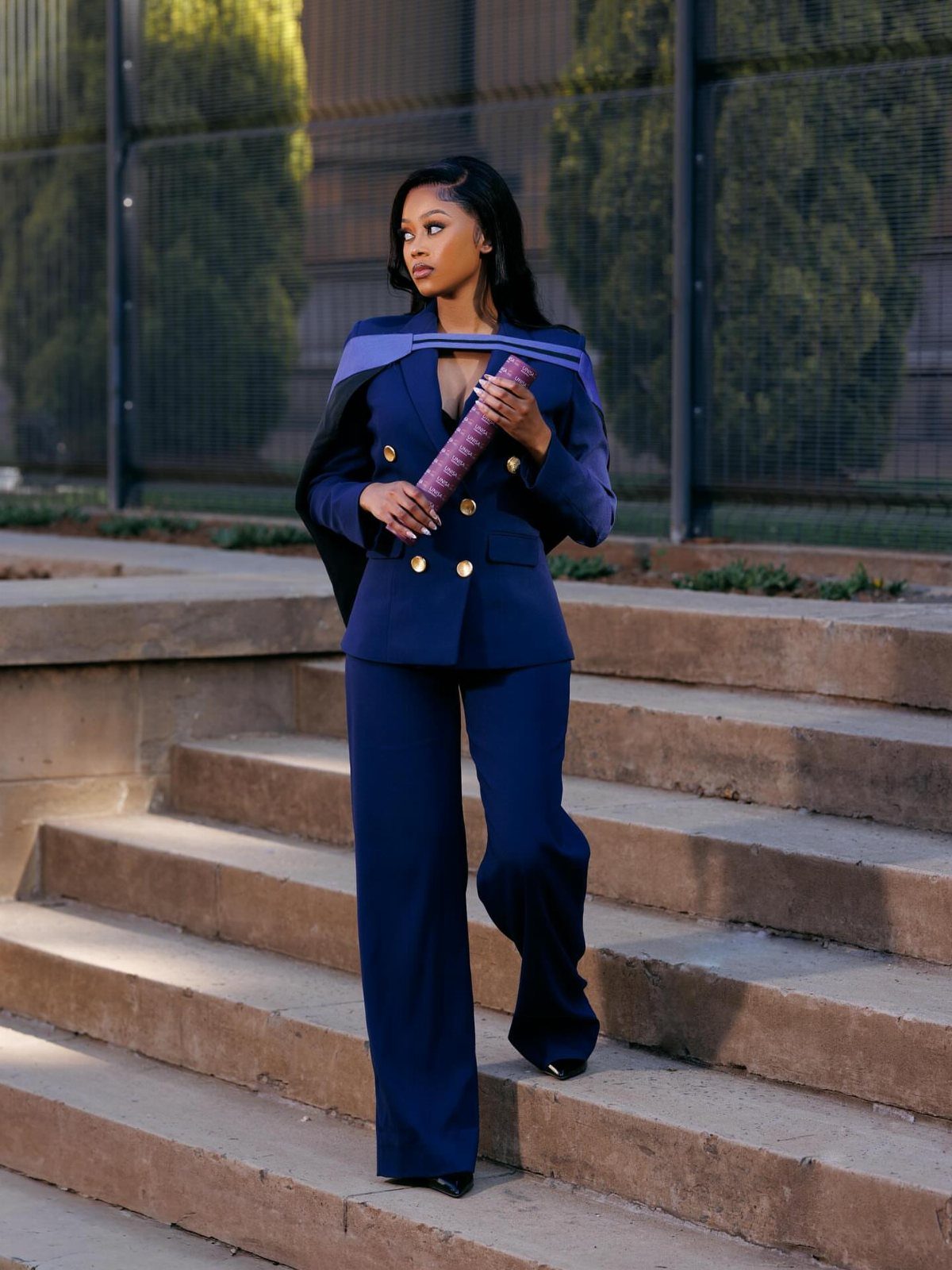 Woman in navy blue pantsuit holding a purple diploma on concrete steps.