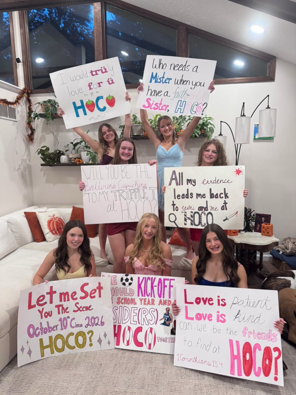 Six teenage girls holding various handmade homecoming proposal posters in a living room.