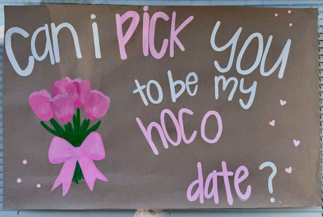 Brown cardboard sign with pink flowers and text asking someone to hoco date