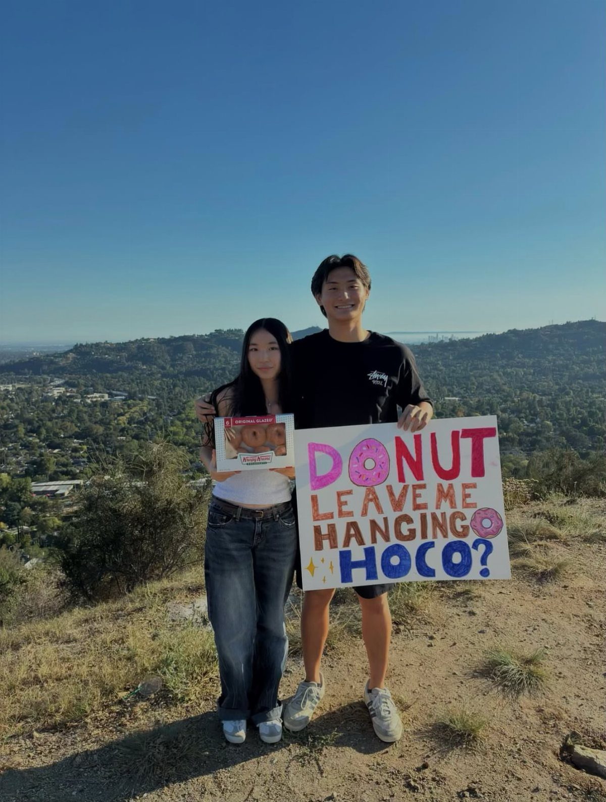 Couple on a hill with a donut themed homecoming proposal poster and donuts