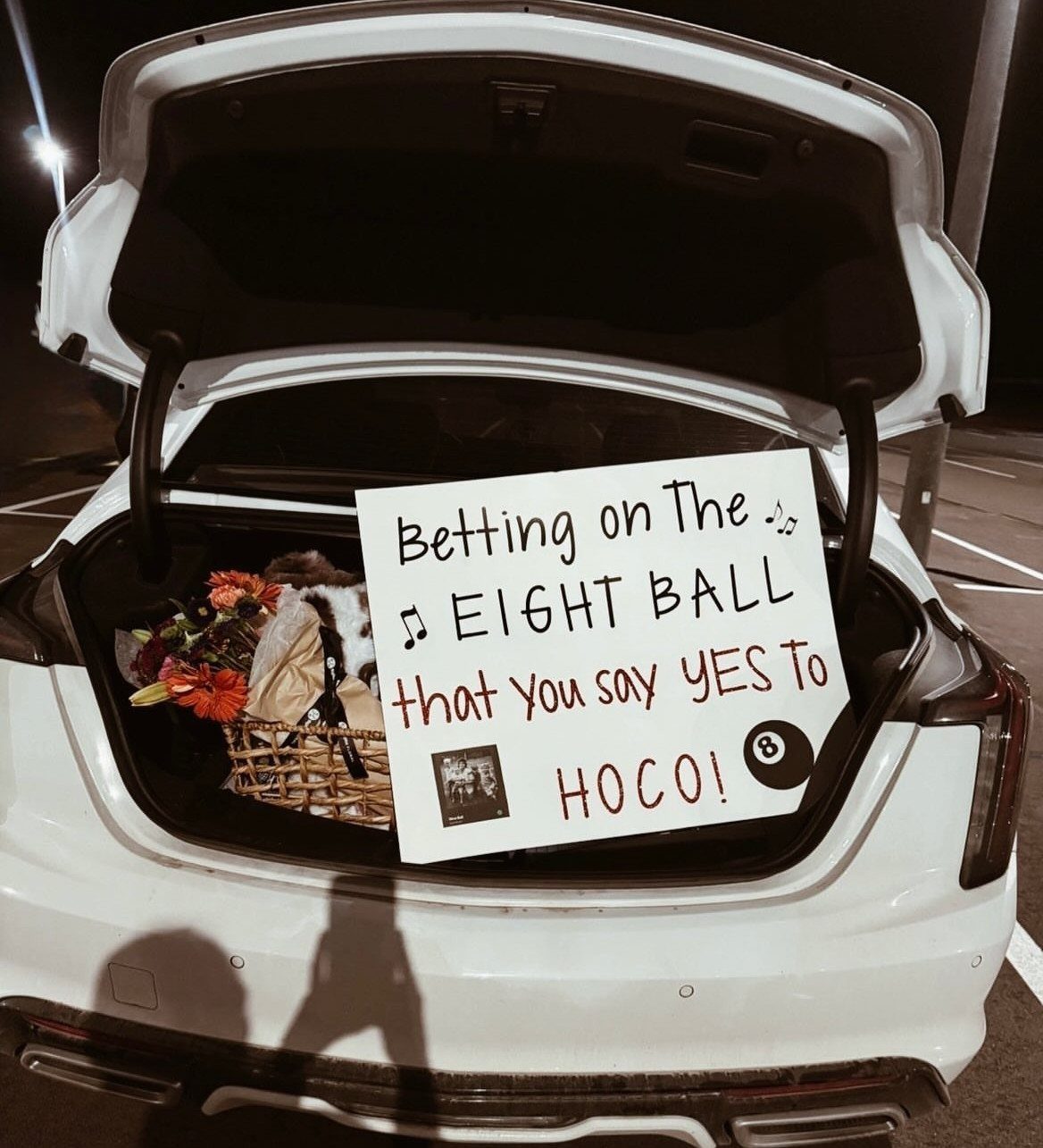 Open white car trunk with a homecoming proposal poster and a flower basket.